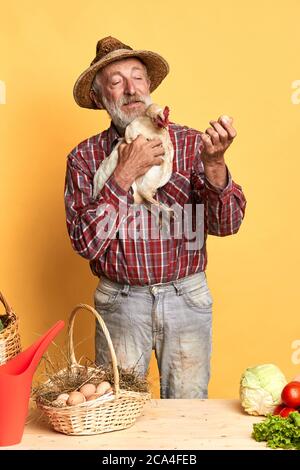 Ritratto verticale dell'uomo del vecchio villaggio in cappello di paglia che tiene il pollo e che guarda alle uova appena posate, andando vendere alcuni al mercato degli agricoltori la settimana Foto Stock