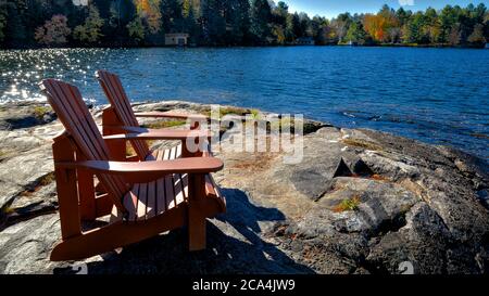 Due sedie Muskoka sedute su una riva rocciosa di fronte ad un lago tranquillo con alberi sullo sfondo Foto Stock