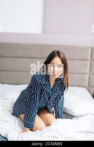 Positivo bella ragazza in camicia di moda a righe seduta a letto e sorridente alla macchina fotografica a casa. Primo piano foto Foto Stock