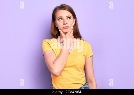 Vista frontale di una giovane donna incerta piena di dubbi, tenendo il mento, guardando a destra pensieroso, si sente esitante, vestita casualmente, in posa su leggero p Foto Stock