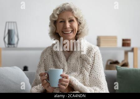 Donna anziana che tiene la tazza di relax sul divano avvolto in plaid Foto Stock