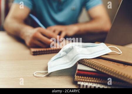 Studentesse maschi a casa durante la quarantena. Primo piano di un tavolo con computer portatile, notebook e maschera medica Foto Stock