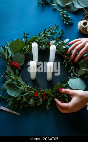 Donna che fa una moderna corona di Natale dell'Avvento con candele bianche, foglie di eucalipto e bacche rosse su sfondo blu. Foto Stock