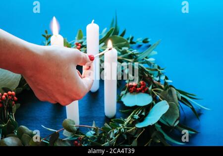 Primo piano di una donna che illumina le candele di Avent su moderno Corona di Natale con foglie di eucalipto isolate su sfondo blu Foto Stock
