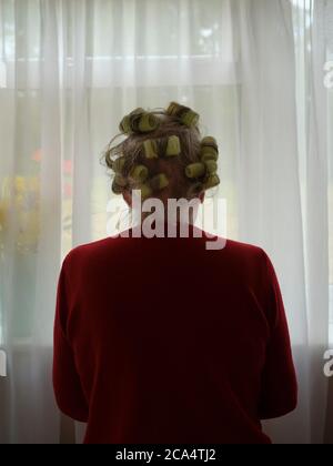 Una donna anziana anziana si autoisola durante il Coronavirus / Covid pandemic lock con i suoi capelli auto lavati e set in rulli guardando la sua finestra. Foto Stock
