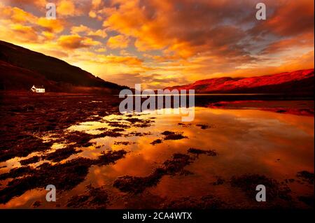 Un paesaggio impressionante con il sole dorato che si riflette in un Scozzese Loch e mettendo in risalto le montagne su Horizon.A cottage bianco è anche visibile Foto Stock