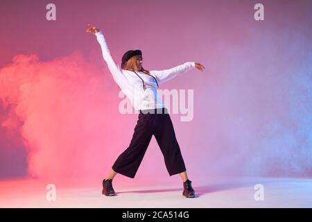 Giovane ragazza allegra con lunghi capelli rossi, con pantaloni neri, pullover sportivo bianco e cappello elegante, danza energica funk dance, isolato in colore Foto Stock