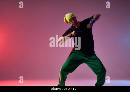 Un uomo impressionante e entusiasta con divertente berretto giallo, pantaloni verde scuro e t-shirt nera, che esprime elementi hip hop a tutto il corpo, godendosi ballare, isolarsi Foto Stock