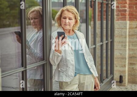 Anziana caucasica graziosa donna di affari circa 62 anni con il telefono mobile in mano sta leggendo il messaggio con il viso serio. Indossa la moda Foto Stock