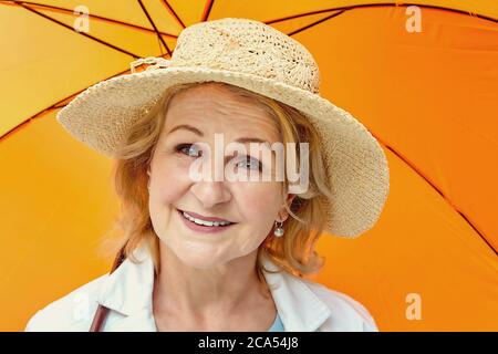 Senior bianco attraente donna bionda circa 62 anni in cappello è in piedi sotto ombrello arancione durante il giorno. È felice, positiva e amichevole. Foto Stock