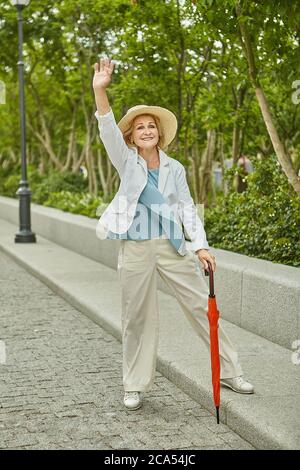 Invecchiato bianco caucasico bella donna in viaggio in tessuto casual è in piedi vicino alla strada e agitando una mano e sorridendo. È attiva e allegra. Foto Stock