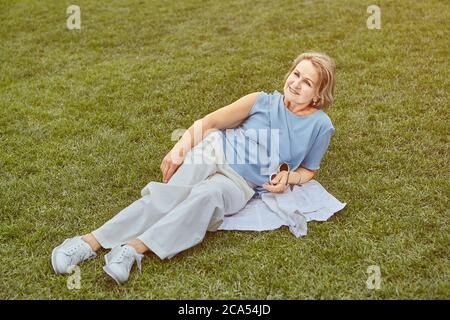 Anziana donna caucasica attiva circa 62 anni è sdraiata sull'erba nel parco pubblico. Indossa abiti eleganti casual. La signora anziana ha un riposo, Foto Stock