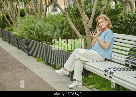 Anziana bella donna bianca di circa 62 anni è seduta sulla panchina nel parco pubblico e guardando nel suo cellulare. Sembra elegante nel suo clo casual Foto Stock