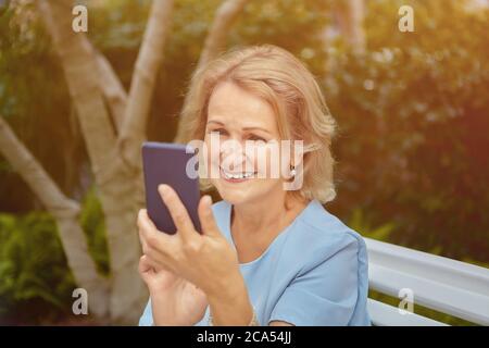 Anziano caucasica bella donna circa 60 anni sta guardando il suo smartphone con sorriso e felice faccia. È seduta sulla panchina nel parco pubblico. Foto Stock