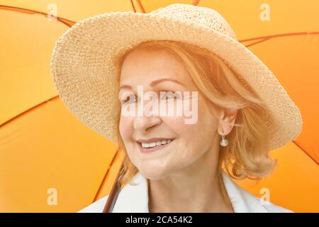 Anziana caucasica bella donna circa 60 anni in cappello sta sorridendo mentre si sta in piedi sotto ombrello arancione all'aperto durante il giorno. È attiva ed hea Foto Stock