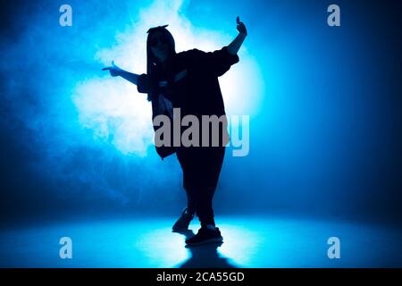 Silhouette giovane donna vestita in stile Street fashion, indossa uno stile hip-hop danzante su uno sfondo blu chiaro da studio con effetti flare Foto Stock