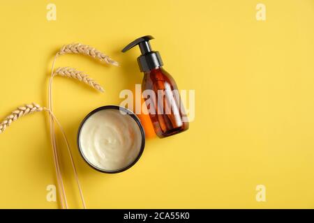 Flacone cosmetico con pompa in vetro ambrato scuro, crema idratante in vaso, grano su sfondo giallo. PRODOTTI DI bellezza naturali BIOLOGICI SPA, concetto di cura della pelle. Foto Stock