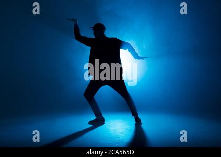 Una silhouette di uomo che offre performance da solo, ballando da solo in stile hip hop sulla scena del club con fulmini al neon blu e fumo. Foto Stock
