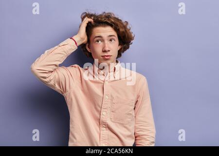 Giovane uomo pensieroso con fracche e capelli lunghi ondulati, indossa una camicia casual a manica lunga di colore chiaro, guarda in su con espressione confusa, fissa curly h Foto Stock