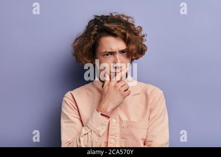 Ritratto di broccia espressiva da uomo adolescente che si maglia, guardando la macchina fotografica, in piedi con uno sguardo pensivo, mettendo la mano sul mento, esprimendo il pensiero Foto Stock