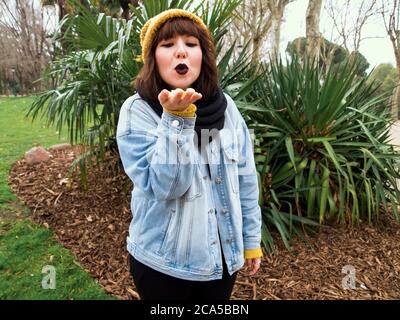 Giovane donna con cappuccio giallo in lana in piedi e baci soffiati. Concetto urbano autunnale. Foto Stock