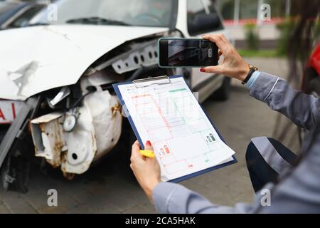 L'agente assicurativo scatta le foto dell'automobile schiantata sul suo smartphone e compila l'assicurazione Foto Stock