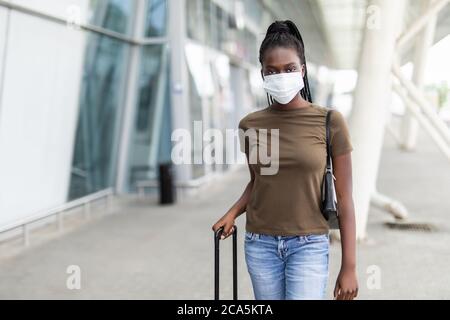 Donna africana è seduta in aeroporto con un computer portatile in una maschera medica. La giovane ragazza sta aspettando la partenza al cancello, lavorando online. Respo sociale Foto Stock