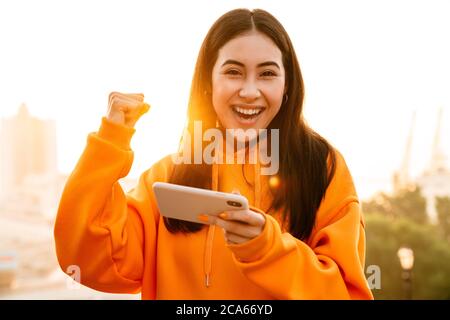 Foto di una donna asiatica entusiasta che fa il gesto del vincitore mentre si gioca al videogioco sul cellulare all'aperto Foto Stock