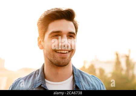 Foto di un gioioso uomo non rasato in una camicia in denim che sorride e guarda da parte mentre cammini all'aperto Foto Stock