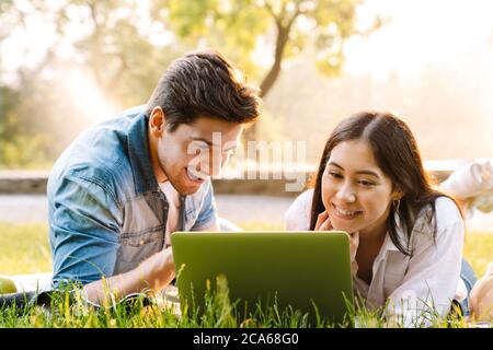 Immagine di una gioiosa coppia multiculturale che usa un computer portatile e sorride mentre si sdraia sull'erba nel parco Foto Stock