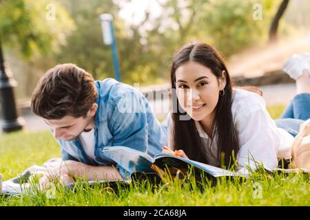 Immagine di una gioiosa coppia studentesca multiculturale che fa i compiti e sorride mentre giace sull'erba nel parco Foto Stock