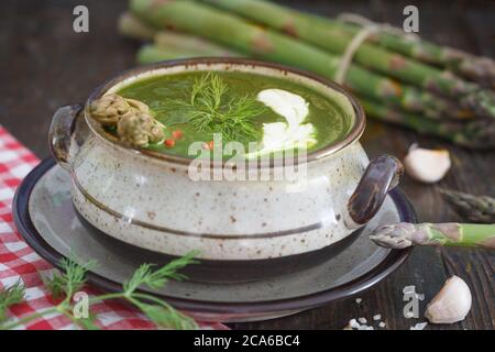 Zuppa di asparagi cremosa decorata con crema, aneto e pepe rosso. Asparagi verdi sullo sfondo, tavolo in legno. Foto Stock