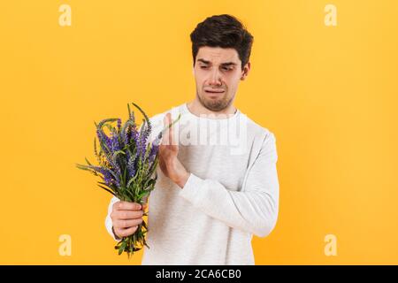 Foto di bell'uomo infelice con allergia in posa con fiori isolati su sfondo giallo Foto Stock