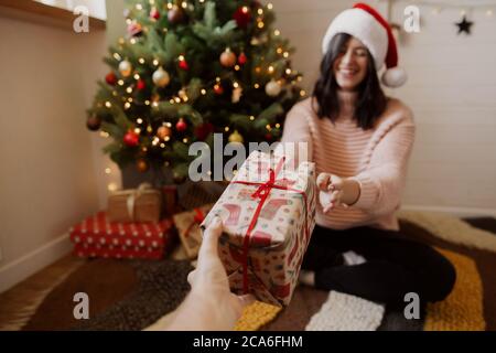 Giovane donna di stile che riceve scatola regalo di natale sotto l'albero di natale con le luci nella stanza moderna. Felice ragazza in cappello di santa tenuta regalo di natale. H Foto Stock