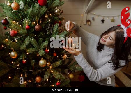 Elegante giovane donna in antlers renne che decorano l'albero di natale con luccicante bauble in camera moderna. Felice ragazza che si prepara per le vacanze invernali, arrangiare Foto Stock