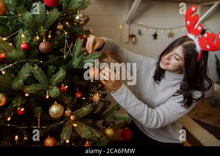 Elegante giovane donna che decora l'albero di natale con luccicante oro bauble in camera moderna. Ragazza felice in antlers renna che si prepara per le vacanze invernali, un Foto Stock