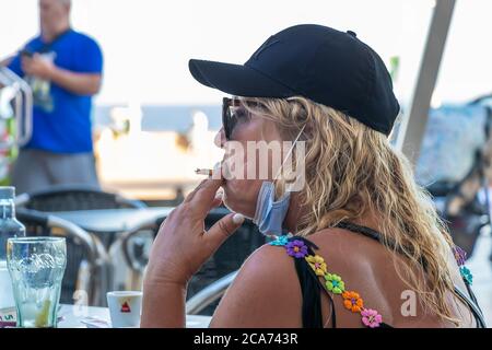 Huelva, Spagna - 10 luglio 2020: Donna bionda matura che indossa la maschera facciale per evitare il coronavirus covid-19 è fumare una sigaretta in una terrazza bar Foto Stock