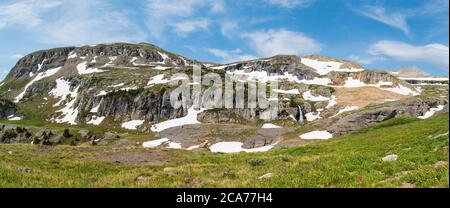 Panorama alpino con montagne innevate, una cascata e un prato alpino con fiori selvatici viola e gialli Foto Stock