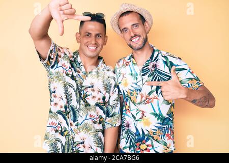 Giovane coppia gay di due uomini con cappello estivo e camicia hawaiana sorridente facendo cornice con mani e dita con viso felice. Creatività e photogra Foto Stock