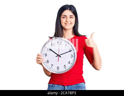 Giovane bella ragazza tenendo orologio sorridente felice e positivo, pollice su facendo eccellente e segno di approvazione Foto Stock