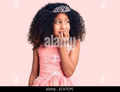 Bambino afroamericano con capelli ricci che indossano la corona della principessa cercando stressato e nervoso con le mani sulla bocca che mordicchiano i chiodi. Problema di ansia. Foto Stock
