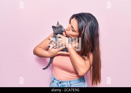 Giovane bella donna latina che puzzava felice. In piedi con sorriso sulla faccia che tiene e baciare adorabile gatto su isolato sfondo rosa Foto Stock