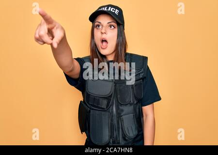 Giovane bella donna di polizia brunette che indossa la polizia uniforme bulletproof e cappuccio puntando con il dito sorpreso davanti, bocca aperta stupito espressione, così Foto Stock