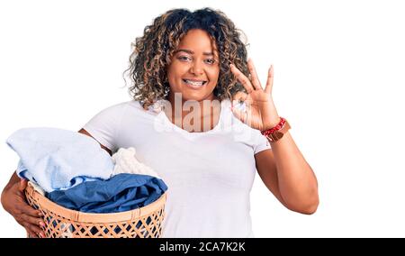 Giovane afroamericano più taglia donna che tiene cesto di bucato facendo segno ok con le dita, sorridente amichevole gesturing simbolo eccellente Foto Stock
