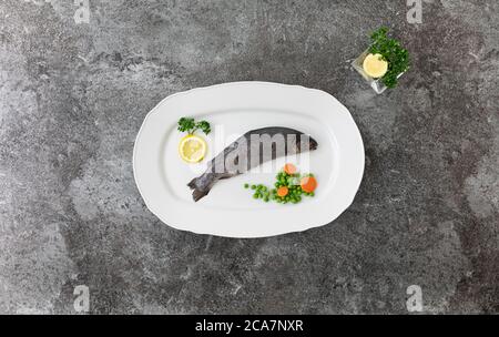 trota iridea intera su un piatto di servizio bianco con limoni, prezzemolo, piselli e carote su un tavolo di marmo grigio Foto Stock