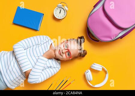 Ritorno a scuola. Una bambina carina con gli occhiali giace su uno sfondo giallo. Libro per bambini sveglia zaino con spazzola cuffie. Concetto di istruzione. In alto Foto Stock