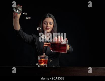 Una ragazza barista attraente versa una bevanda alcolica in un bicchiere e getta pezzi di ghiaccio. Bella brunetta in nero preparare cocktail. Spazio di copia. Foto Stock
