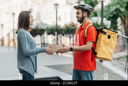 Donna prende l'acquisto in linea dal corriere. Deliveryman in casco, con zaino dà scatola al cliente Foto Stock