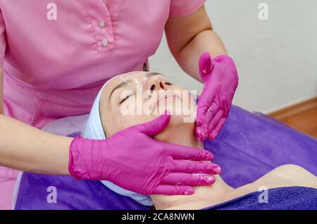 Il medico cosmetologo applica la crema al viso della donna nel salone di bellezza. Preparazione per la procedura di ringiovanimento, restauro della pelle, muscoli facciali Foto Stock