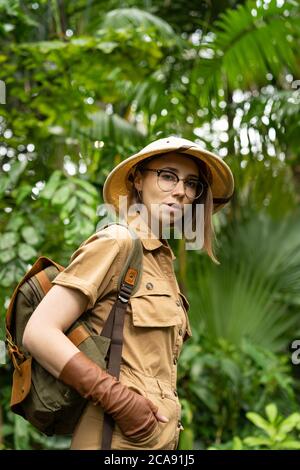 Donna botanica vestita in stile safari in serra. Naturalista in abiti kaki, guanti di lievito con zaino passeggiate nella foresta pluviale circondata da p. Foto Stock